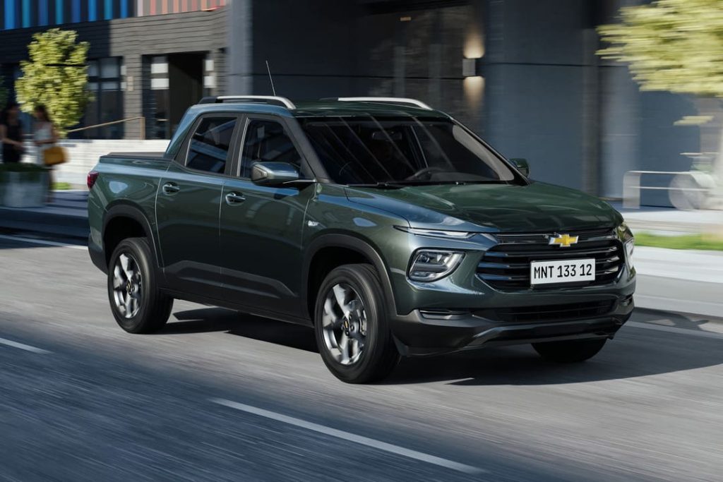 Nova Chevrolet Montana verde escura em movimento em avenida urbana com edifícios ao fundo.