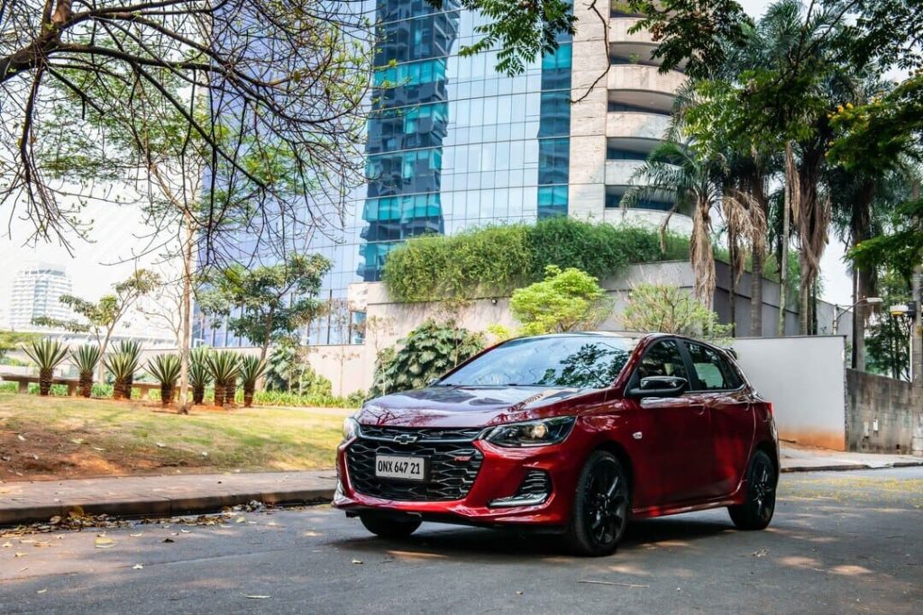 Chevrolet Onix vermelho estacionado em uma rua arborizada de área urbana moderna.
