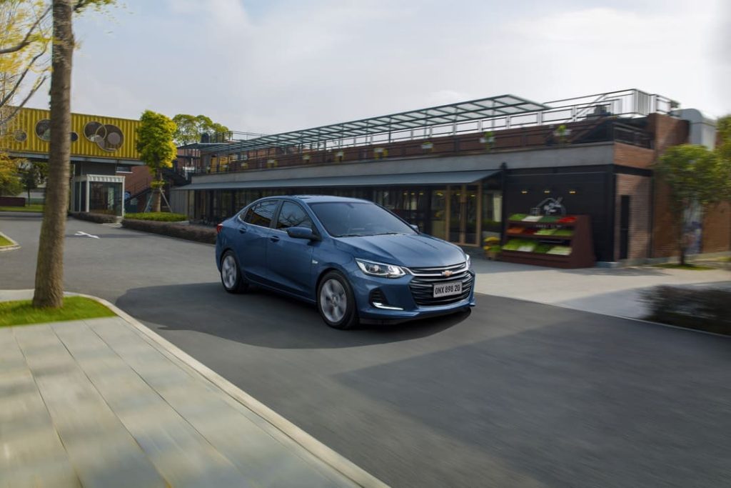 Chevrolet Onix Plus azul circulando em uma rua moderna com paisagismo e prédios comerciais ao fundo.