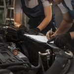 A imagem mostra dois mecânicos realizando uma inspeção em um veículo. Eles estão ao lado do motor do carro, com um deles escrevendo em uma prancheta enquanto o outro observa. Ambos usam uniformes de trabalho e luvas pretas, indicando um ambiente profissional de oficina mecânica. O capô do carro está aberto, revelando partes do motor, sugerindo que estão fazendo uma vistoria ou revisão técnica no veículo.