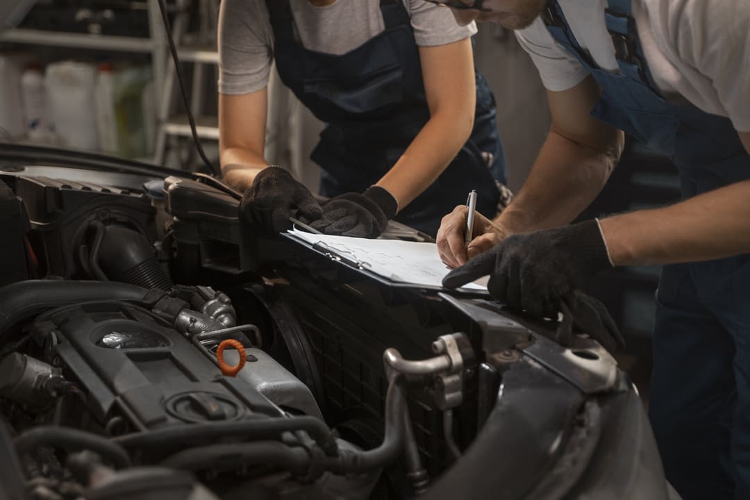 A imagem mostra dois mecânicos realizando uma inspeção em um veículo. Eles estão ao lado do motor do carro, com um deles escrevendo em uma prancheta enquanto o outro observa. Ambos usam uniformes de trabalho e luvas pretas, indicando um ambiente profissional de oficina mecânica. O capô do carro está aberto, revelando partes do motor, sugerindo que estão fazendo uma vistoria ou revisão técnica no veículo.