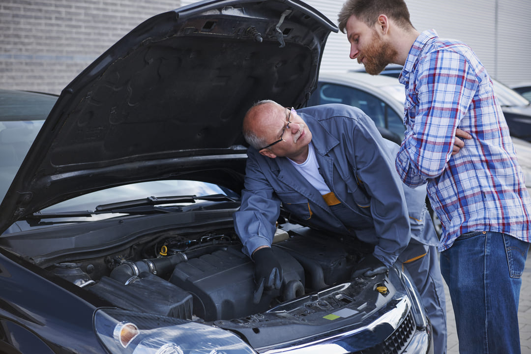 Imagem de um mecânico e um cliente ao lado de um carro com o capô aberto. O mecânico, que aparenta ser mais velho e usa uniforme de trabalho cinza, está olhando para o motor enquanto conversa com o cliente. O cliente, de camisa xadrez e barba, observa atentamente com os braços cruzados, demonstrando interesse e possivelmente preocupação com o que está sendo explicado sobre o veículo.