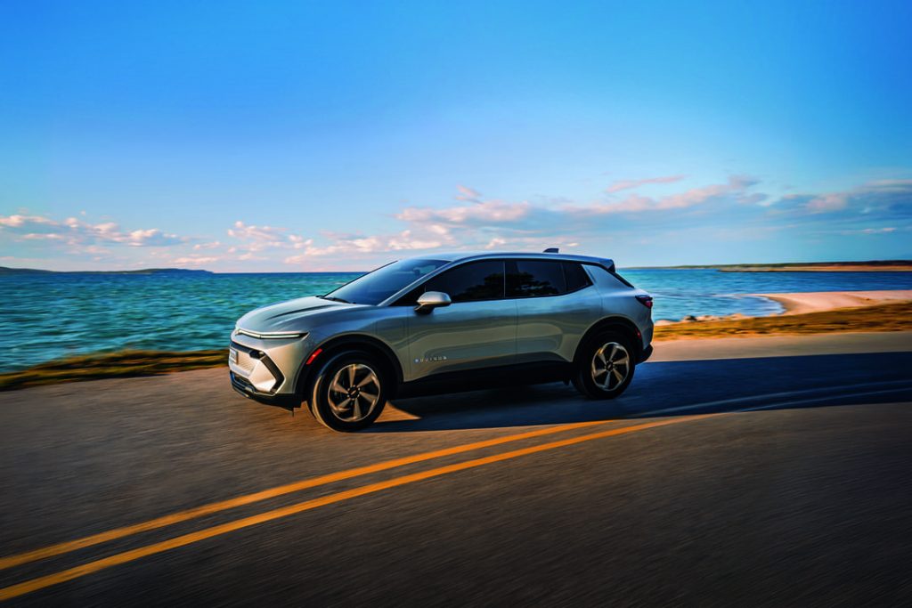 SUV prateado em movimento em uma estrada à beira-mar durante o entardecer, com céu azul e paisagem costeira ao fundo.