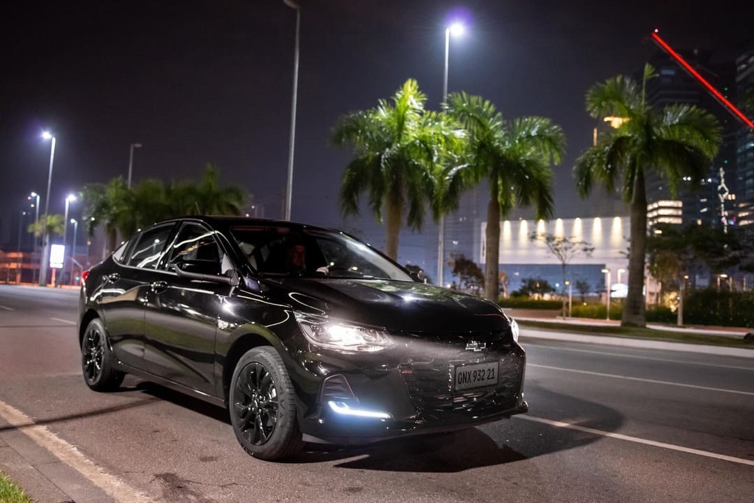 Carro preto com faróis acesos parado em avenida urbana à noite, simbolizando atenção e prática de direção defensiva.
