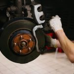 Mecânico com luvas realiza manutenção em sistema de freio a disco de veículo, destacando pinça e disco de freio.