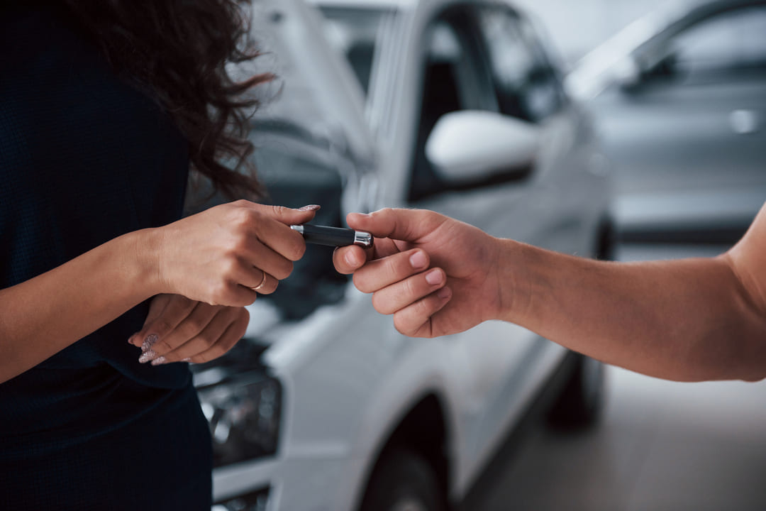 Pessoa entrega a chave de um carro para outra, com um veículo branco ao fundo.