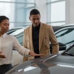 Vendedora apresenta carro a cliente em concessionária, sugerindo o momento de compra ideal.