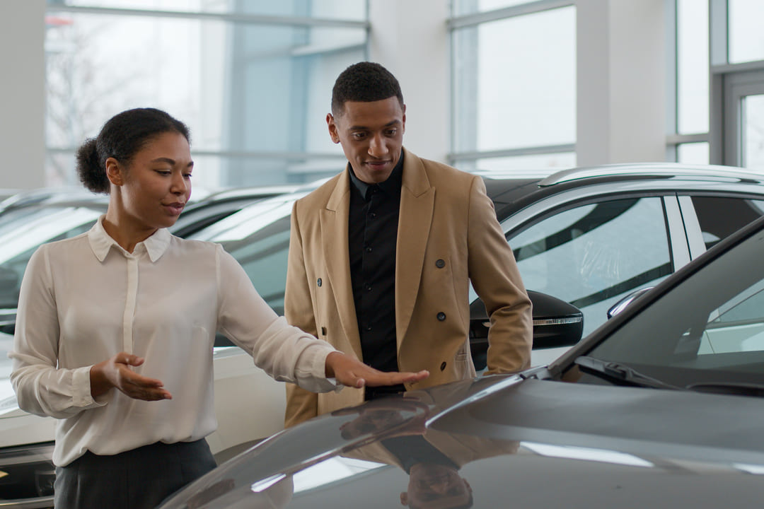 Vendedora apresenta carro a cliente em concessionária, sugerindo o momento de compra ideal.