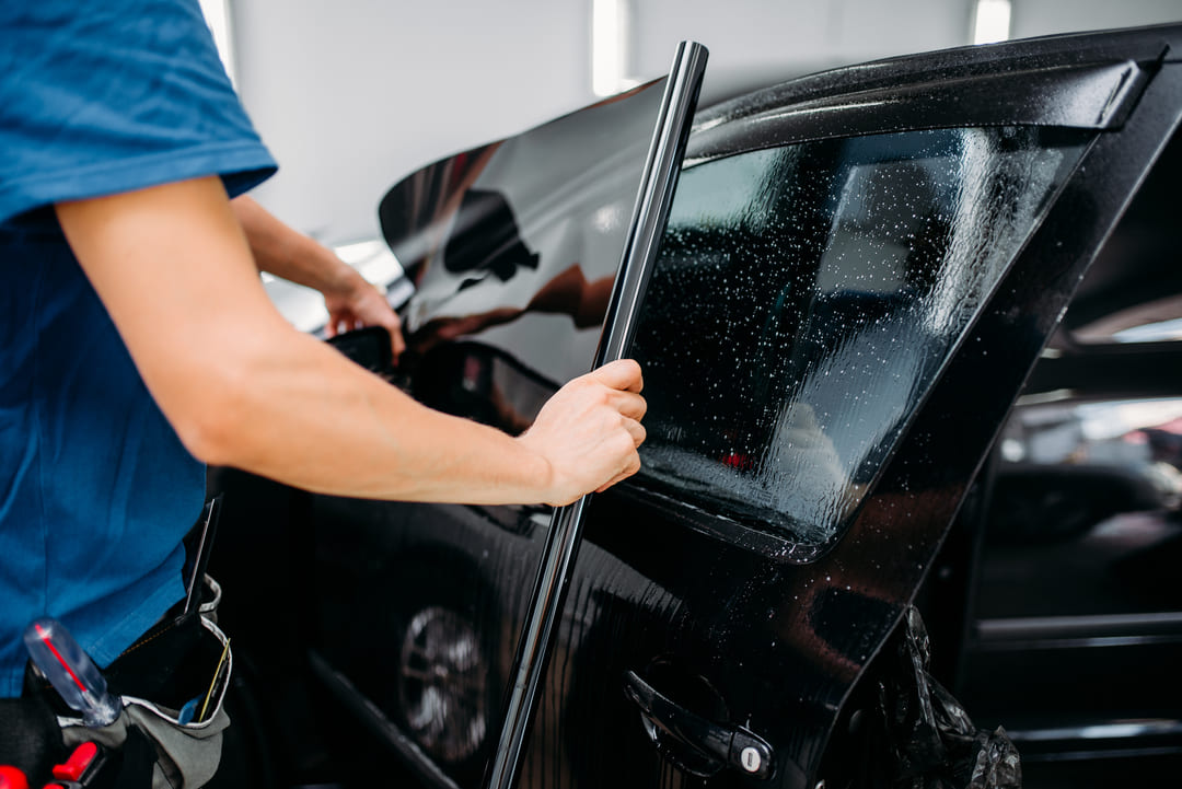 Pessoa aplicando película Insulfilm na janela lateral de um carro preto, com gotas de água visíveis no vidro.