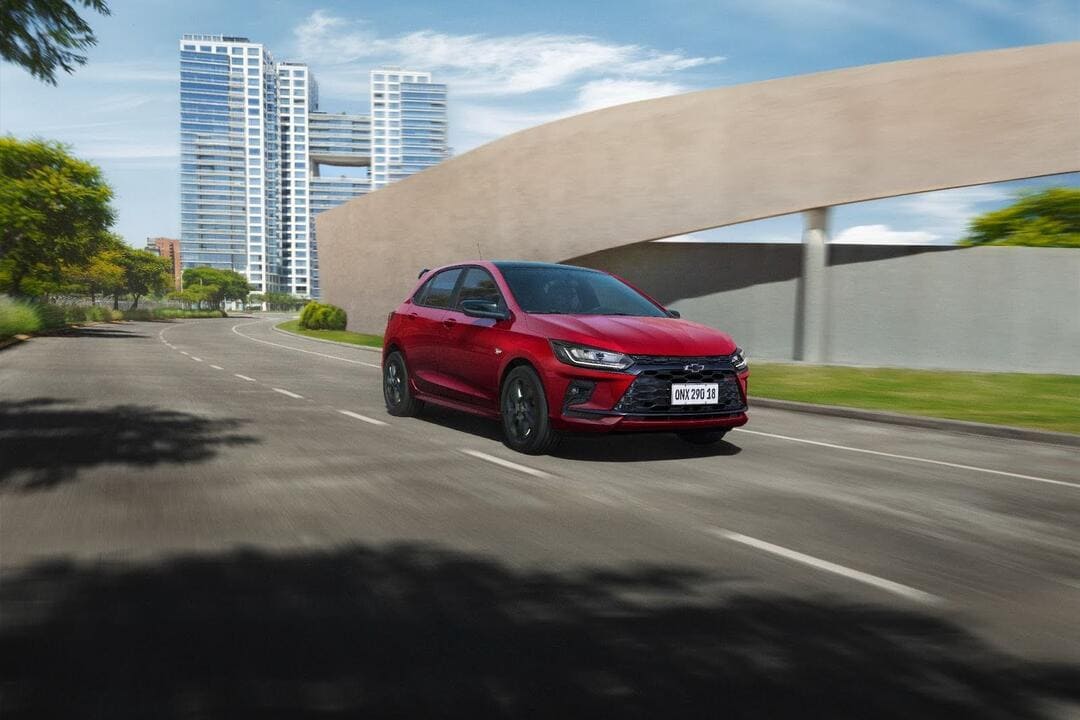 Imagem de um carro vermelho Chevrolet Onix em movimento em uma rua de cidade, com edifícios modernos ao fundo. A imagem está focada no design do carro, destacando sua estética e o cenário urbano.