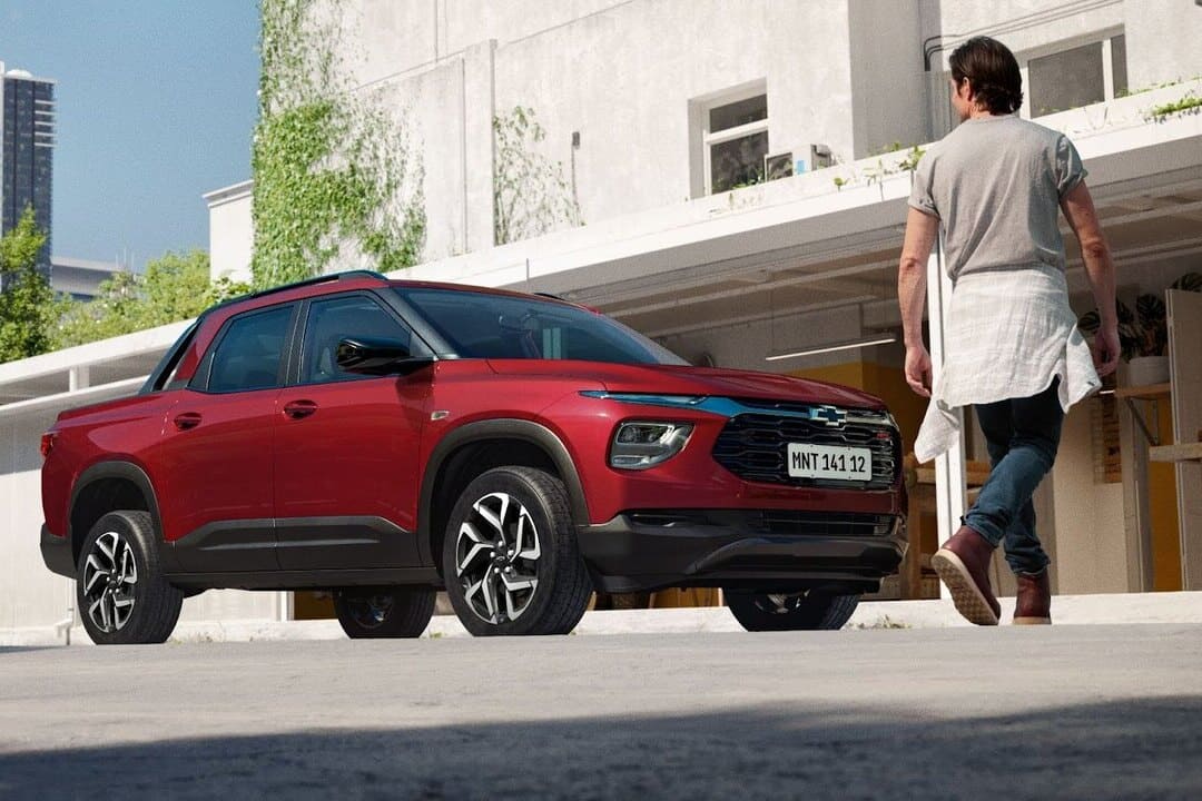 Homem caminhando em direção a uma picape Chevrolet vermelha estacionada em frente a um prédio moderno, representando a possibilidade de adquirir o veículo por meio de financiamento e consórcio Chevrolet.